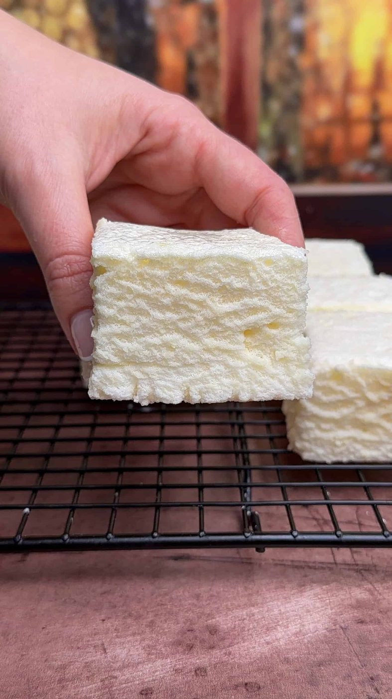 A hand holds a thick, square white marshmallow with a textured surface over a black wire cooling rack, with more marshmallows and a blurred background visible.