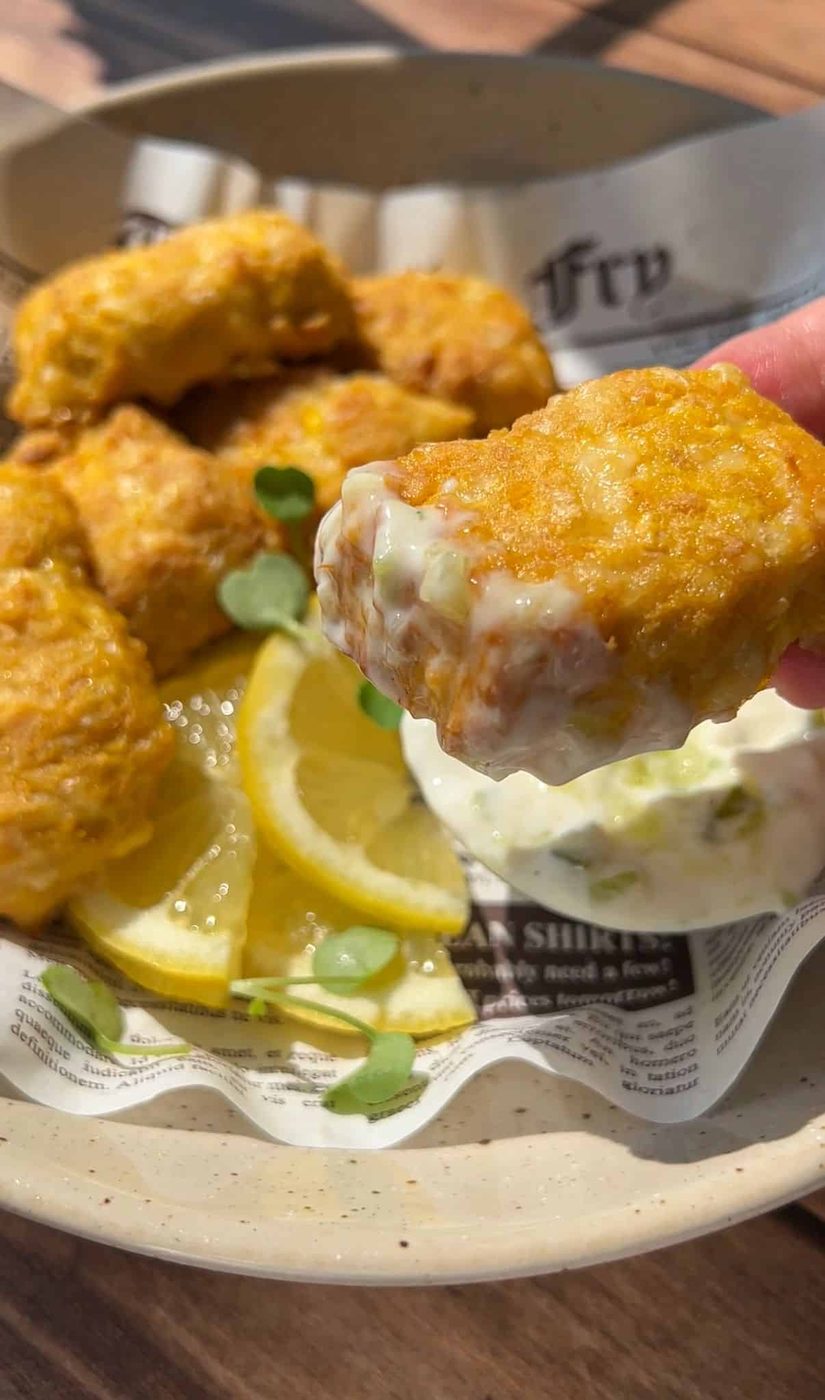 A hand dips a golden, crispy nugget into a creamy white sauce. The plate holds several more nuggets, lemon wedges, and fresh green herbs, all arranged on a sheet of printed paper.