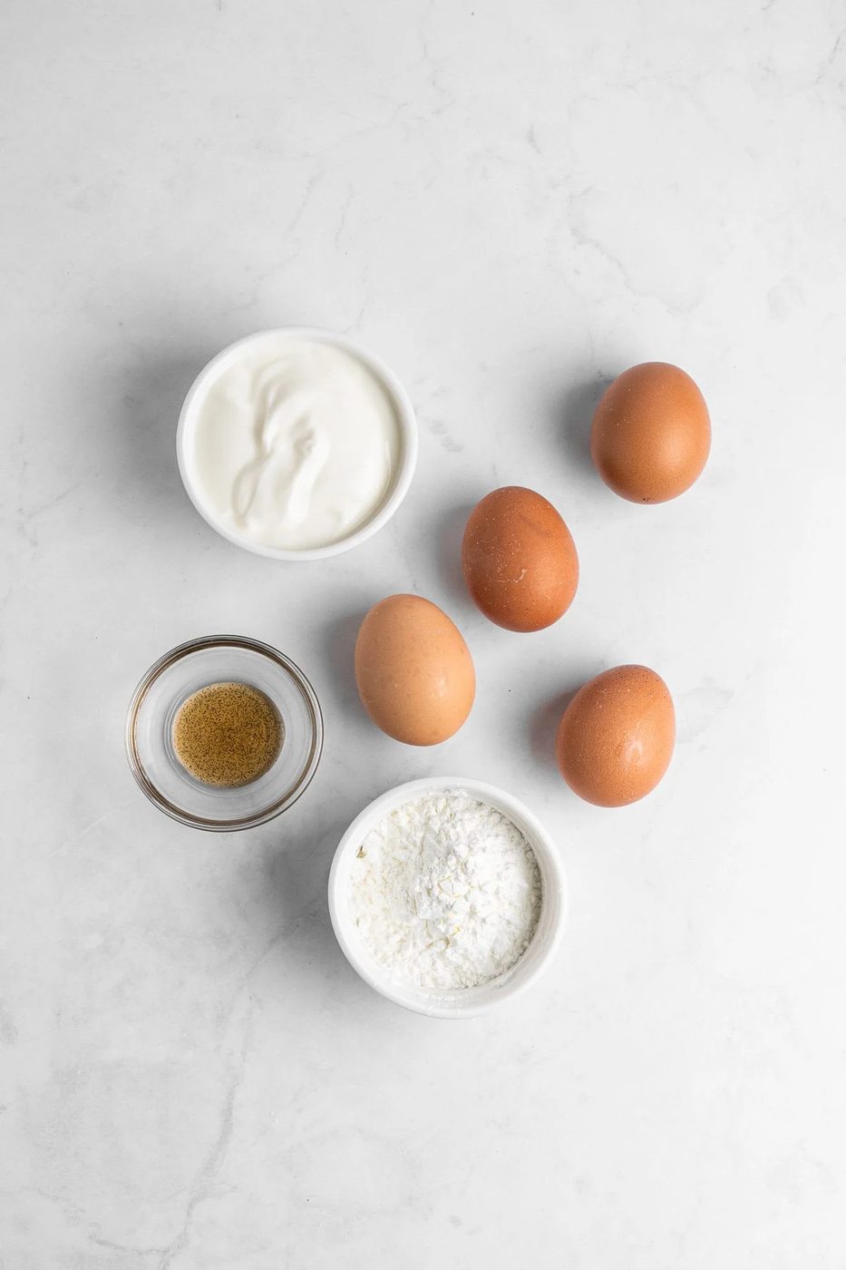Four brown eggs, a small bowl of vanilla extract, a bowl of yogurt, and a bowl of flour are arranged on a white marble surface.