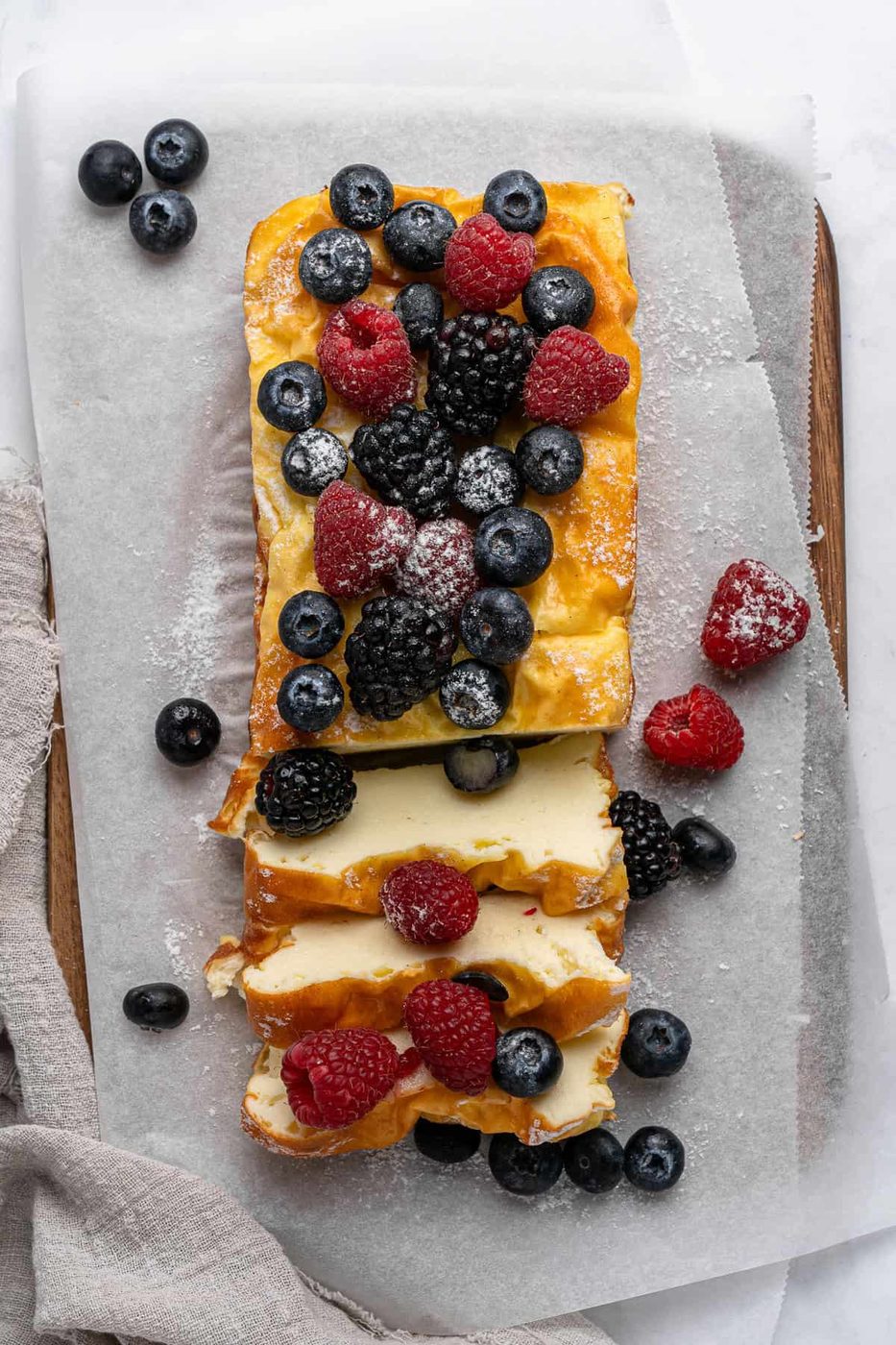 A 3 Ingredient Yogurt Cake topped with fresh blueberries, raspberries, and blackberries rests on parchment paper. Some slices are cut, revealing a creamy inside. A dusting of powdered sugar decorates the berries. A cloth napkin is nearby on the wooden board.