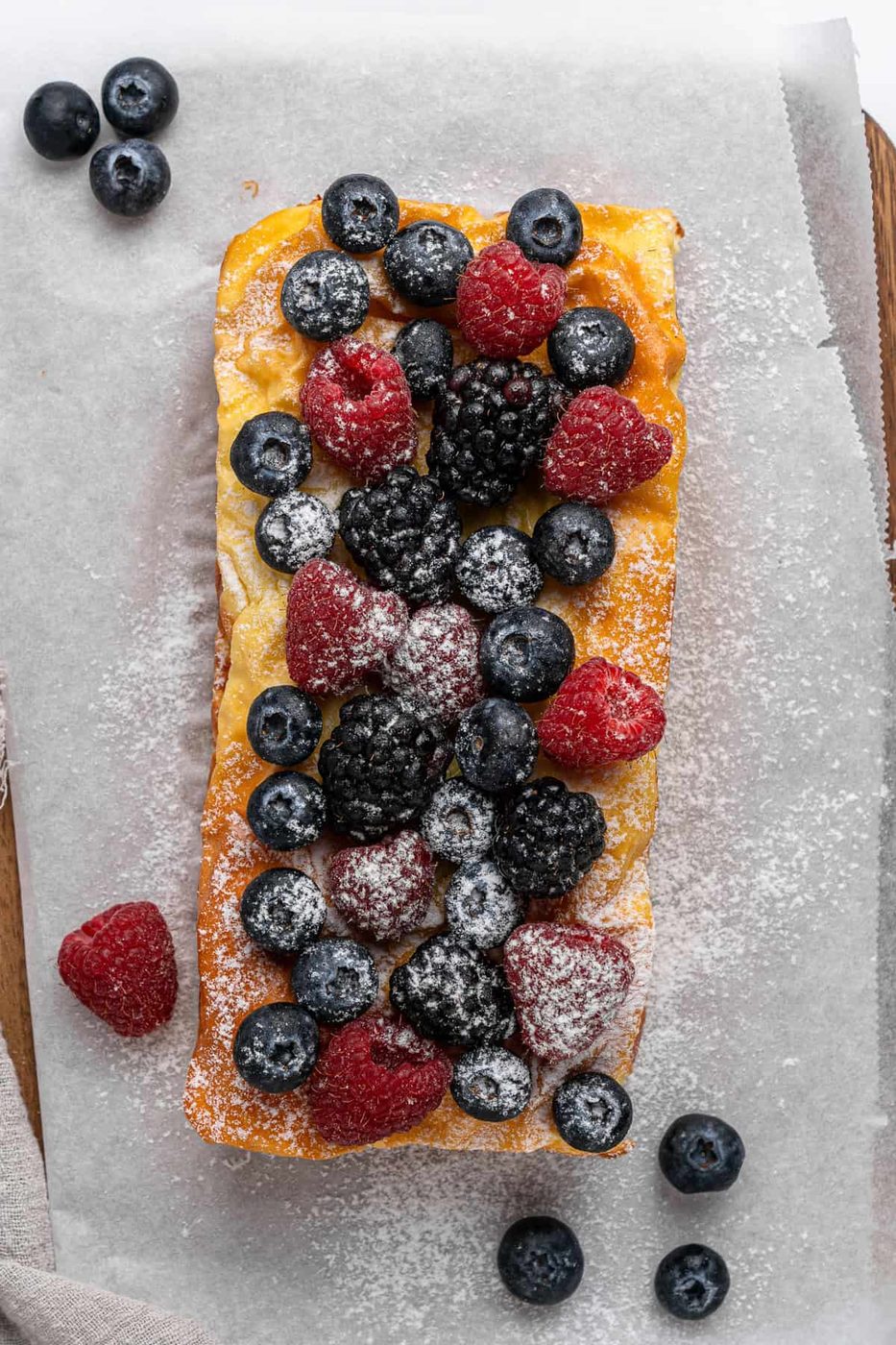 A rectangular A 3 Ingredient Yogurt Cake topped with fresh blueberries, blackberries, and raspberries, dusted with powdered sugar. It's placed on a sheet of parchment paper with a few scattered berries around it.