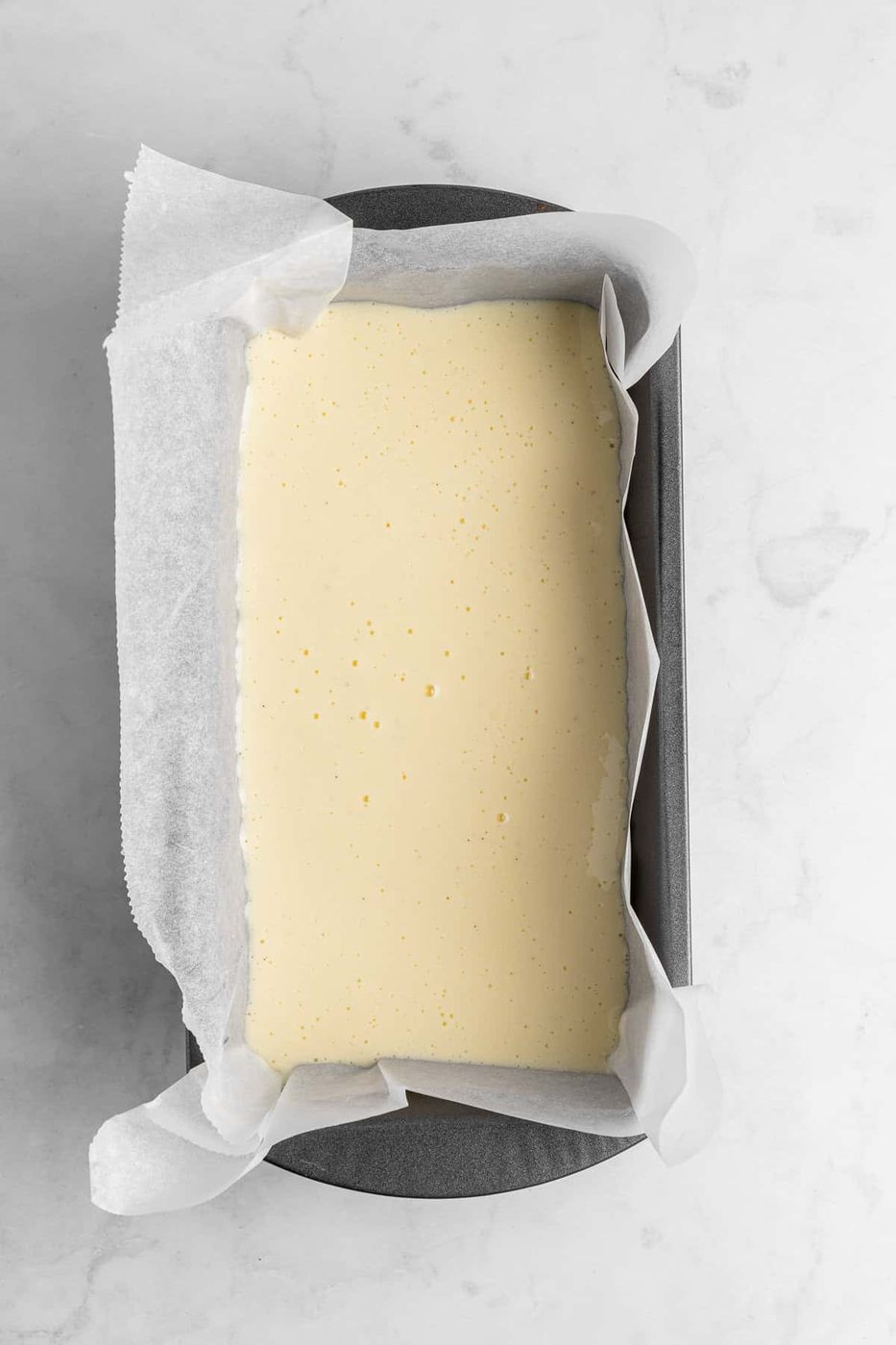 A loaf pan lined with parchment paper is filled with creamy yellow batter, ready to be baked. The pan is set on a light marble countertop.