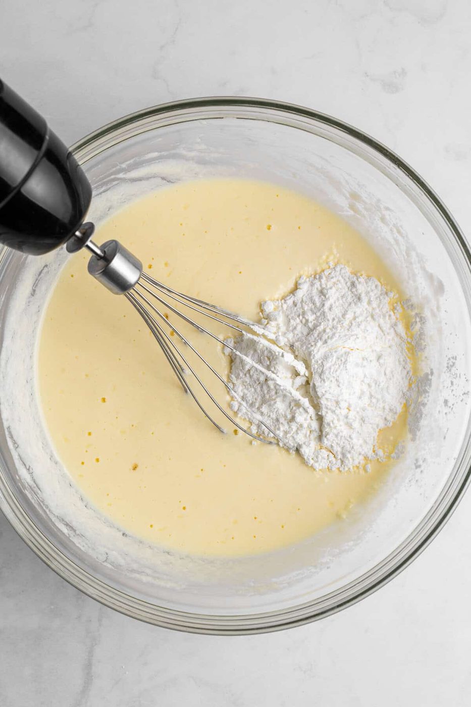 A glass bowl containing cake batter with a heap of flour on one side. An electric hand mixer with a whisk attachment is partially submerged in the batter, ready to mix the ingredients on a marble countertop.