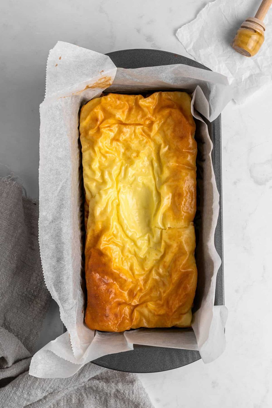 Golden brown, freshly baked bread in a parchment-lined loaf pan on a marble surface. A honey dipper with honey is nearby, along with a grey cloth.