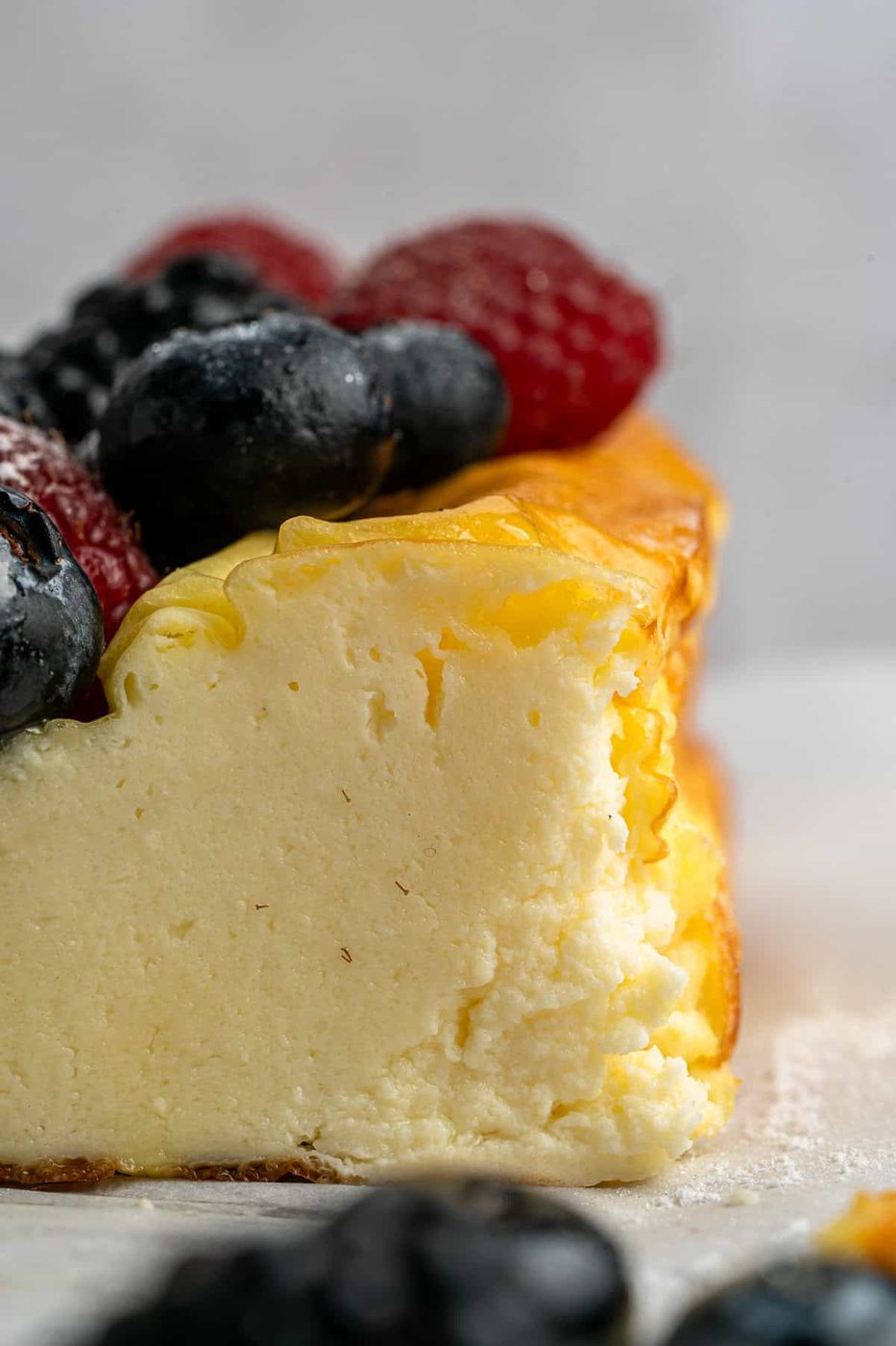 A close-up of a creamy cheesecake with a golden top, topped with fresh blueberries and raspberries. A piece has been cut from the side, revealing its smooth texture. Dusting of powdered sugar is visible.
