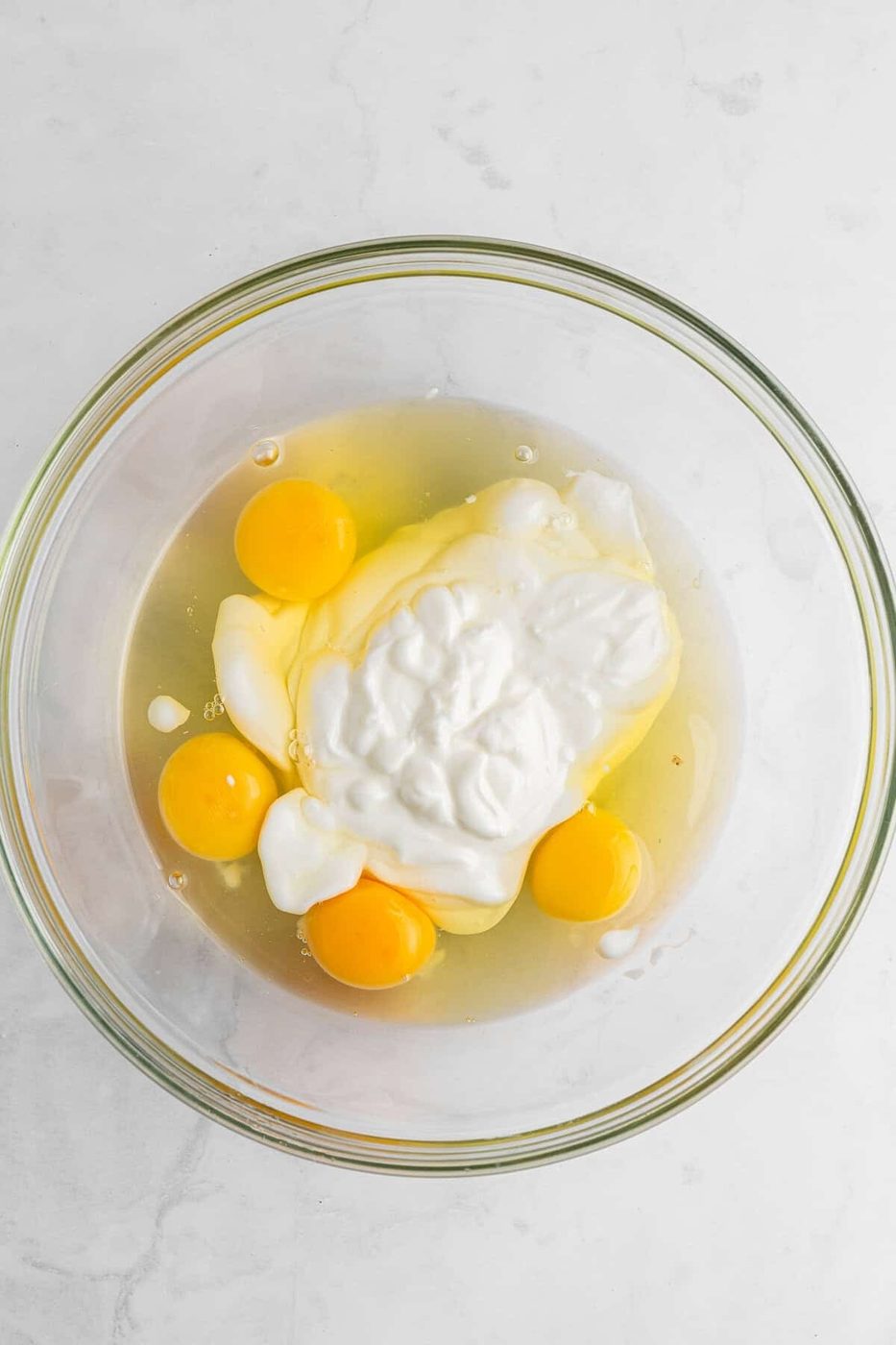A glass bowl containing four cracked eggs and a dollop of yogurt or cream on a white marble surface.