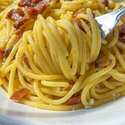 Close-up of a bowl of classic carbonara, with creamy sauce, crispy bacon pieces, and a fork twirling a portion of the pasta.