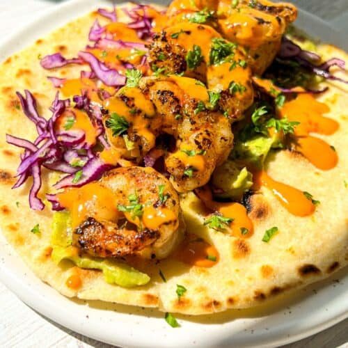 A shrimp taco on a white plate, topped with grilled shrimp, shredded purple cabbage, avocado, fresh herbs, and drizzled with an orange sauce. The taco is on a light wooden table, bathed in bright sunlight.