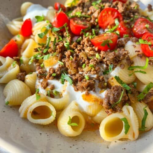 A bowl of pasta with creamy sauce, topped with seasoned ground meat, cherry tomato halves, and fresh parsley. The dish is served in a light-colored bowl with a spoon. Sunlight casts a warm glow over the meal.