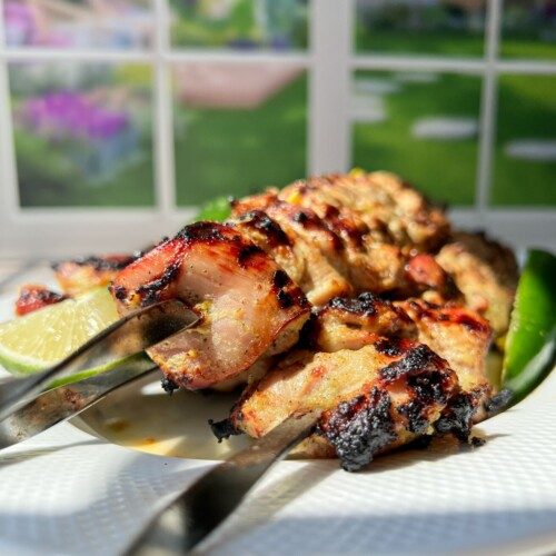 Three metal skewers with grilled chicken kebabs, inspired by Whole Lemon Chicken flavors, are served on a white plate, garnished with a lime wedge and a green chili. Sunlight streams in from a window, revealing a garden view in the background.