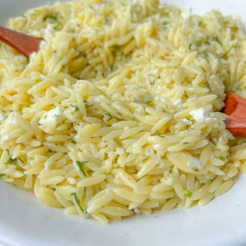 A close-up of a white bowl filled with creamy Lemon Feta Orzo pasta, mixed with herbs and small pieces of cheese, with two wooden utensils resting in the dish.