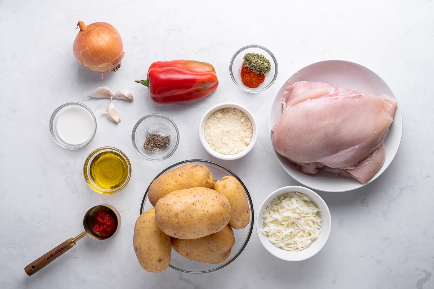 Overhead view of raw ingredients for Chicken and Mashed Potatoes on a white surface: potatoes, chicken breasts, red bell pepper, onion, garlic, shredded cheese, olive oil, milk, spices, and tomato paste in small bowls.