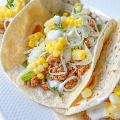 Three soft Ground Beef Tacos filled with seasoned meat, shredded cheese, corn kernels, lettuce, and a drizzle of white sauce, served on a white plate.