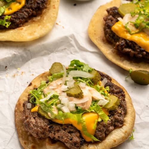Three open-faced cheeseburger flatbreads topped with melted cheese, lettuce, chopped onions, sliced pickles, and sauce, on parchment paper. A small bowl of extra pickles is in the background.