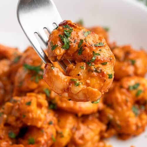 A close-up of a fork holding a piece of cooked shrimp in a spicy red sauce, garnished with herbs. More shrimp covered in the same sauce are visible on a white plate in the background.