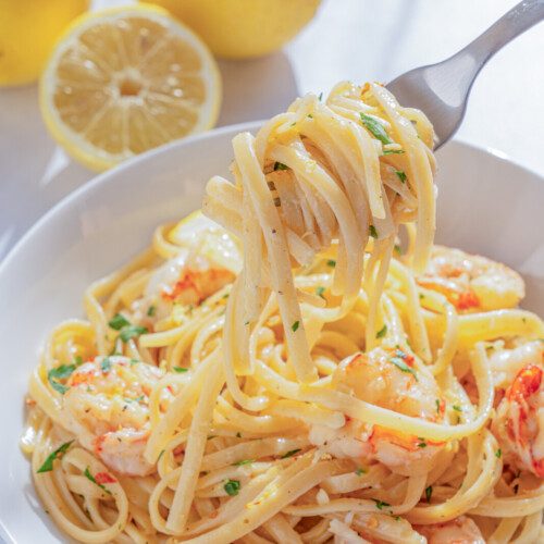 A bowl of Lemon Shrimp Pasta garnished with herbs, with a fork lifting a portion. In the background, two whole lemons and one lemon half rest on a white surface.