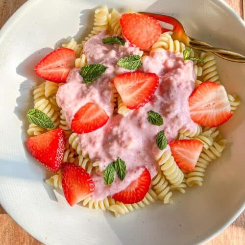 A white bowl of rotini pasta topped with a creamy pink strawberry sauce, fresh strawberry slices, and mint leaves, with a gold fork on the side, on a wooden surface.