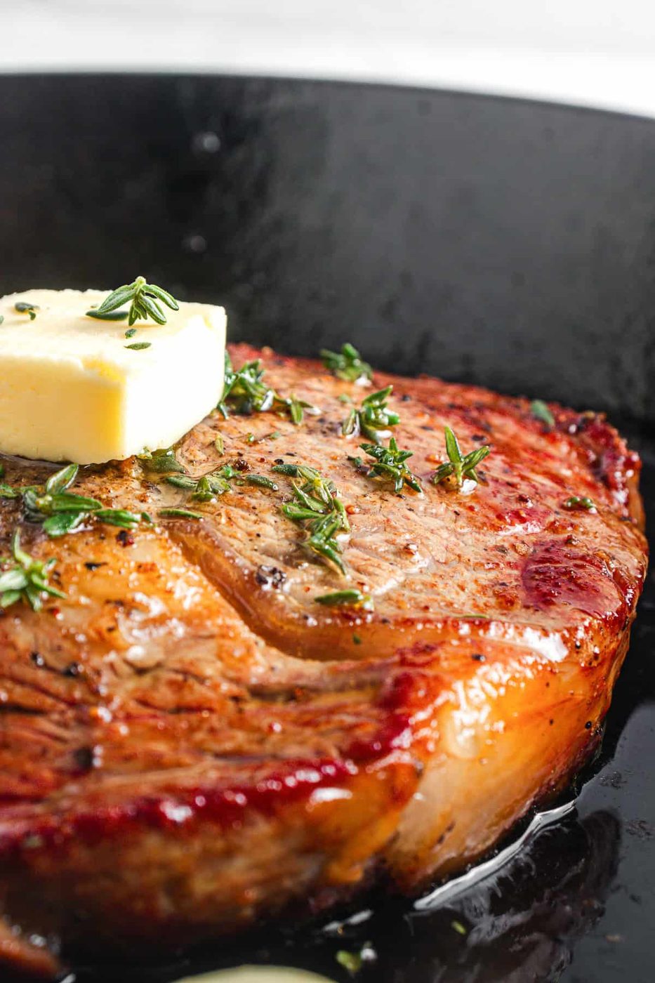 A close-up of a seared steak in a black skillet, topped with a melting pat of butter and sprinkled with fresh thyme. The steak is browned and glistening, with visible grill marks and a juicy appearance.