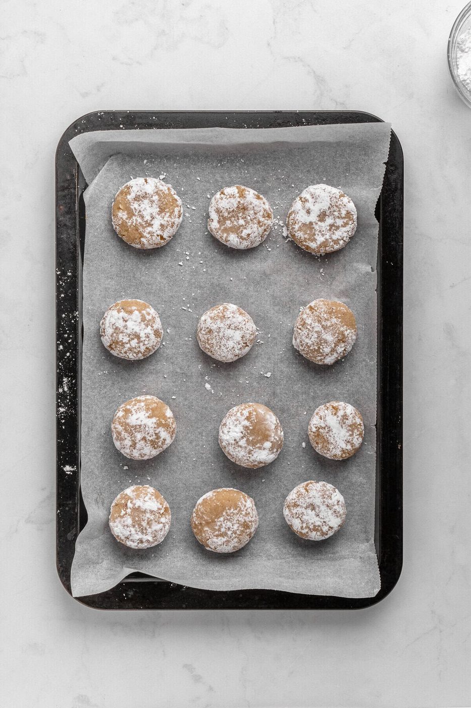 A baking tray with twelve raw, round cookies covered in powdered sugar. The cookies are arranged in four rows on parchment paper, ready for baking. The background is a white, textured surface.