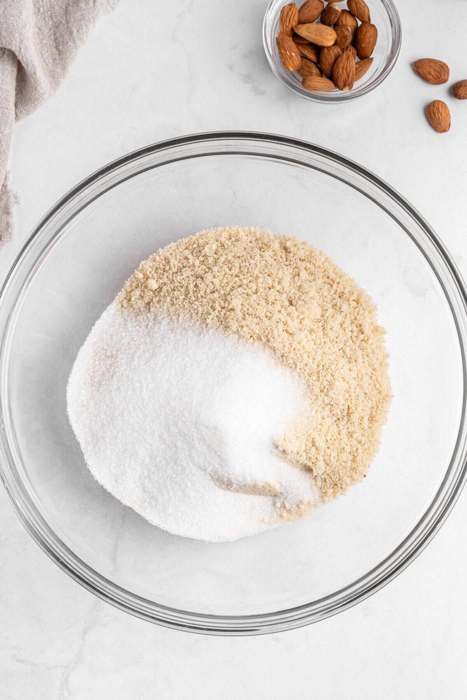 A glass bowl containing white and brown sugar is placed on a marble surface. To the top right, there's a small bowl filled with almonds, and a few almonds are scattered nearby. A beige cloth is partially visible to the top left.
