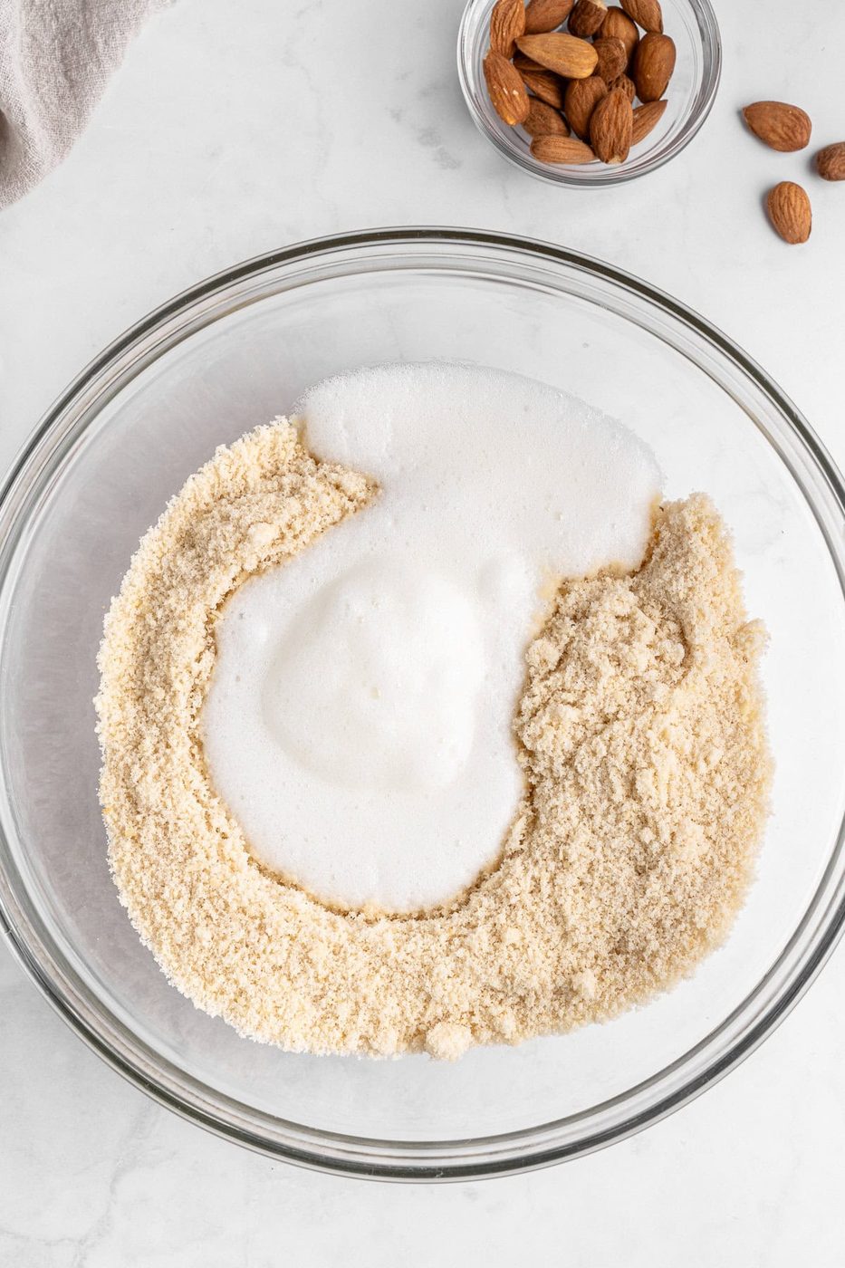 A mixing bowl contains almond flour and foamy egg whites, partially blended together. A small bowl with whole almonds and a few loose almonds are visible in the top right corner on a light-colored marble surface.