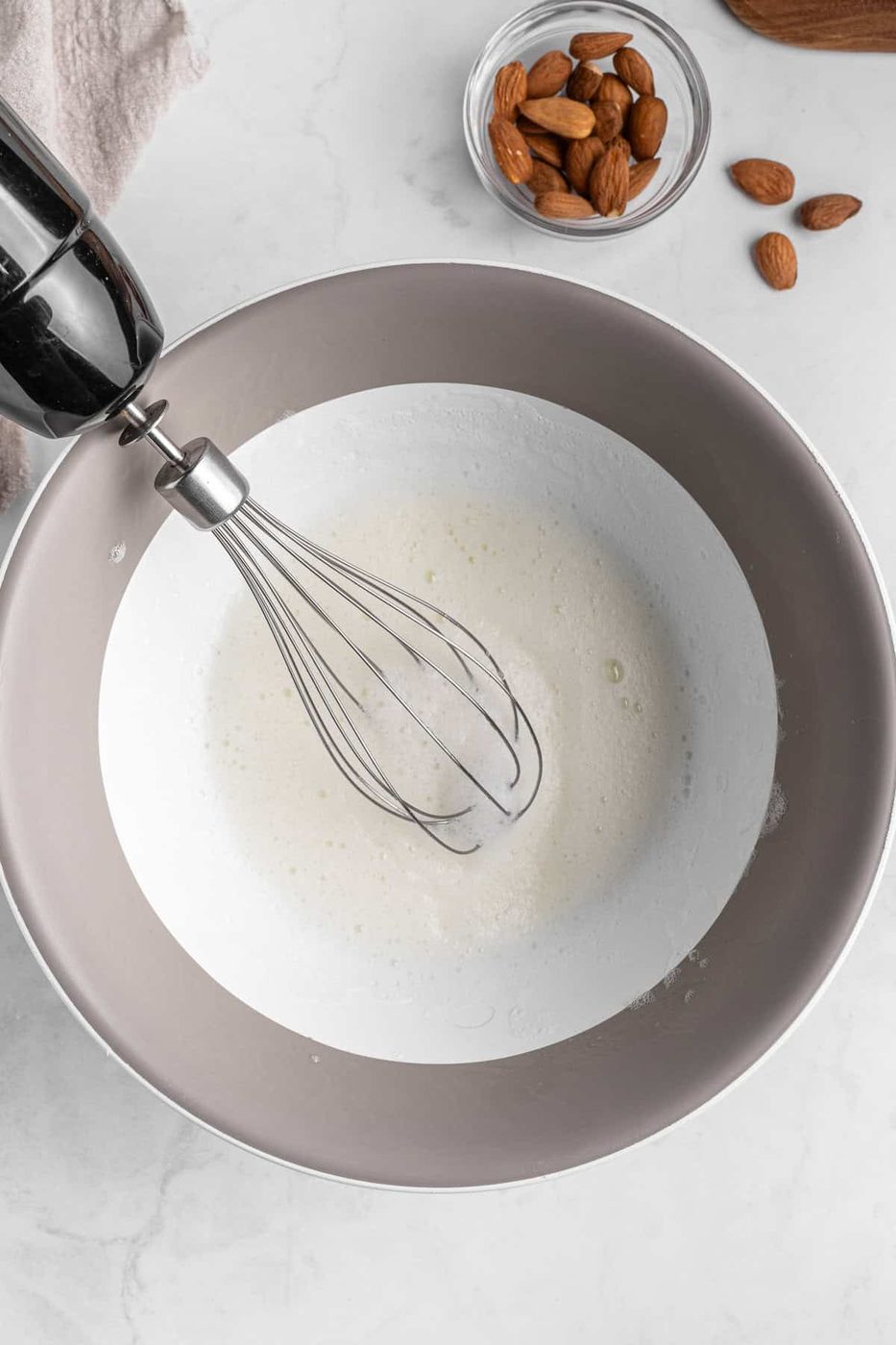 A whisk attached to an electric mixer is beating egg whites in a mixing bowl. Beside the bowl, there is a small glass bowl filled with almonds on a marble countertop.