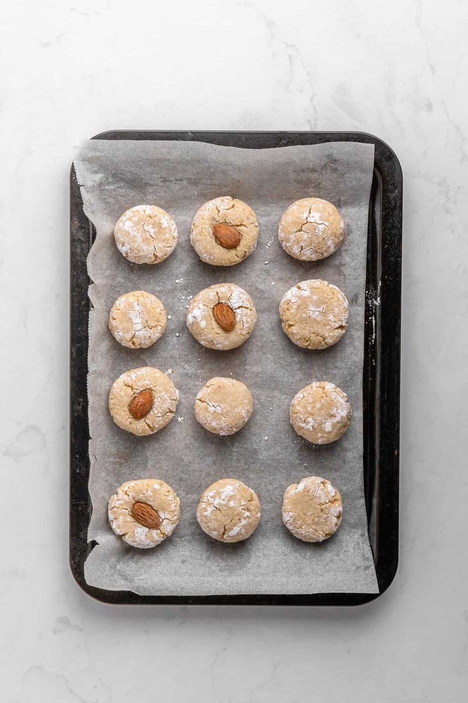 A baking tray lined with parchment paper holds twelve Italian Amaretti Cookies, arranged in a grid. Some cookies have a whole almond on top. The tray rests on a white marble surface.