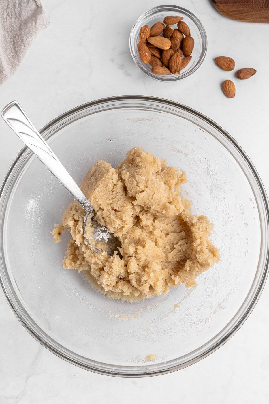 A glass bowl on a marble surface containing mixed almond paste with a spoon in it. A small glass bowl with whole almonds and a few scattered almonds are in the background.