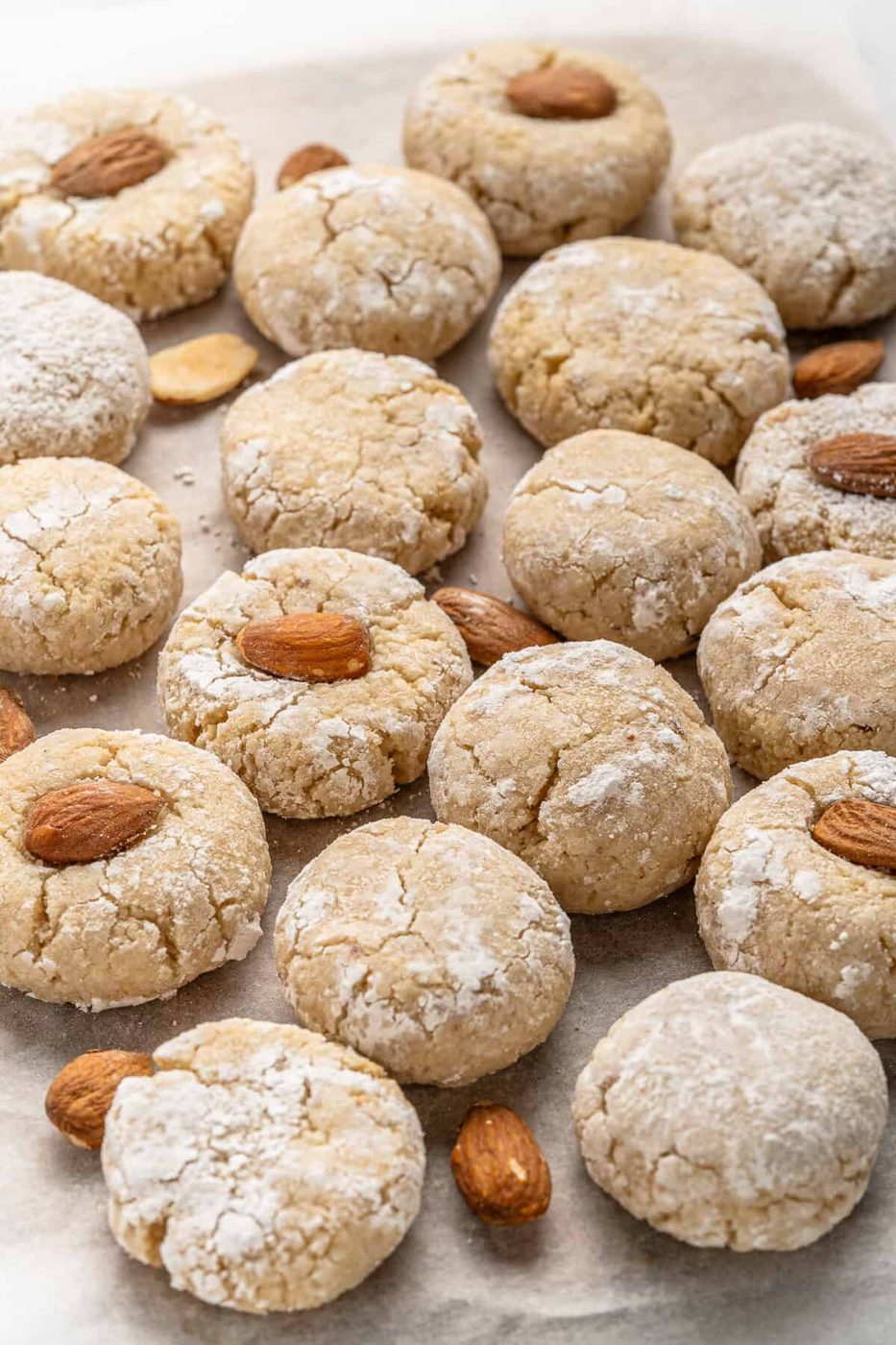 A batch of Italian Amaretti Cookies dusted with powdered sugar is arranged on baking paper. Some cookies have a whole almond on top, while others are plain. A few loose almonds are scattered among the cookies.