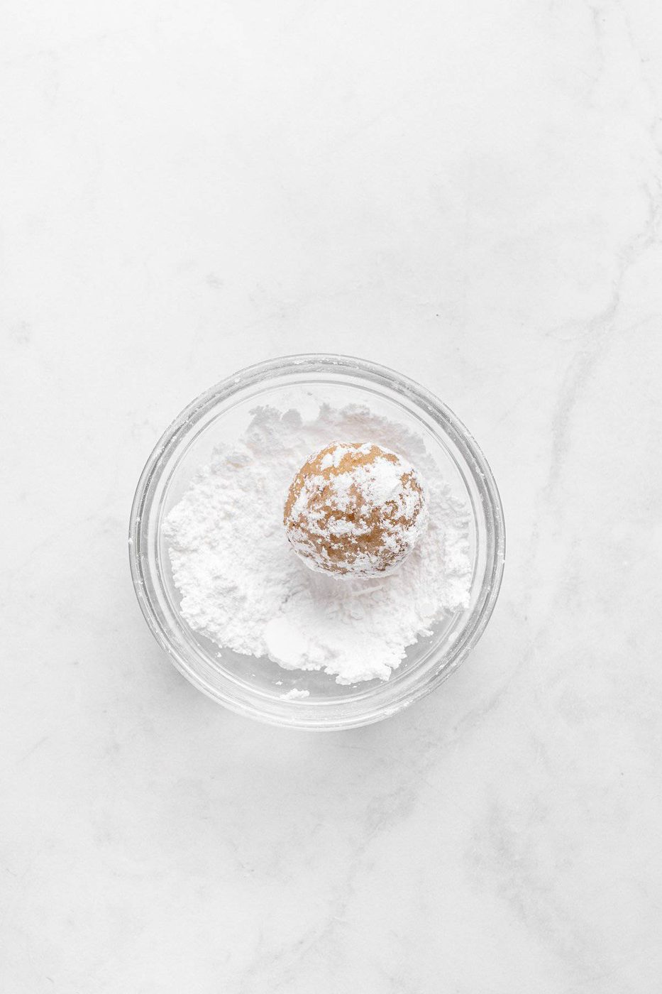 A small round ball of cookie dough sits in a clear glass bowl filled with powdered sugar, placed on a light marble surface.