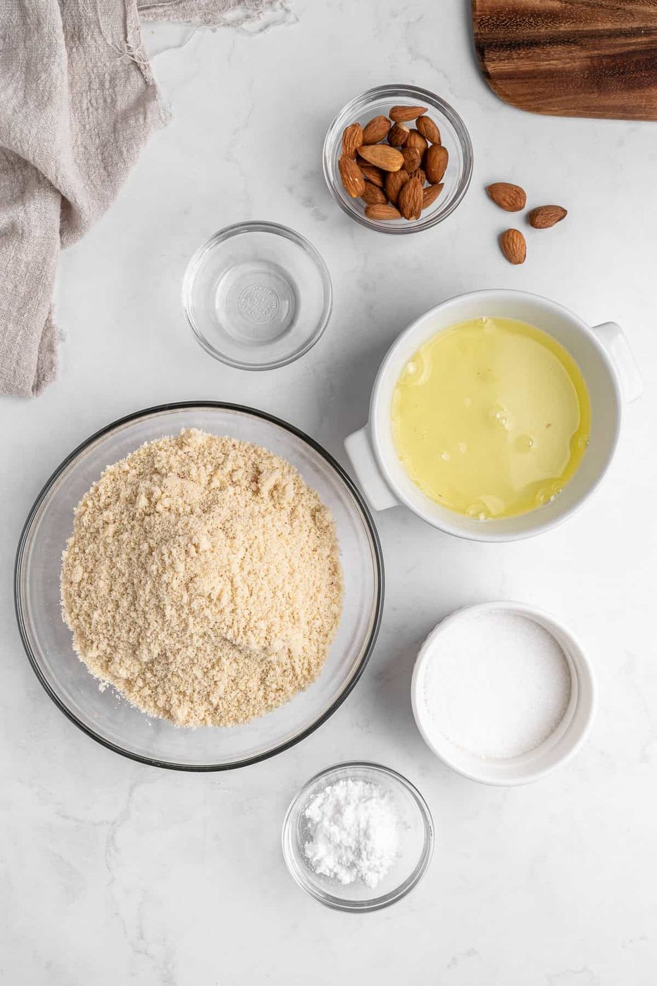 Top-down view of baking ingredients on a white surface, including a bowl of almond flour, a bowl with egg whites, a bowl of whole almonds, a small bowl of powdered sugar, and a glass of water, with a beige cloth nearby.