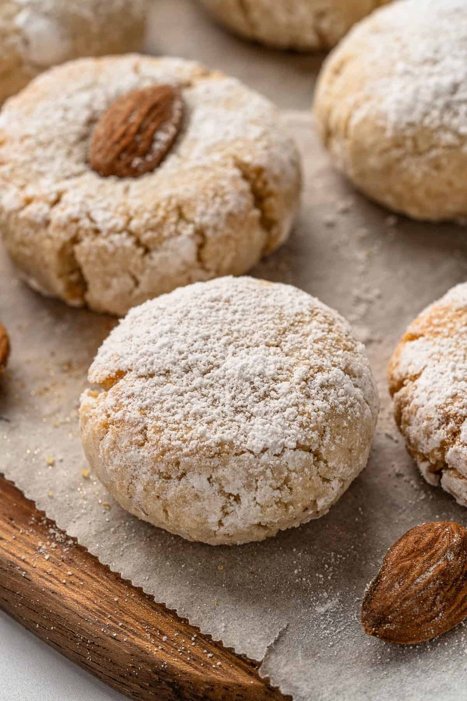 Close-up of freshly baked almond Italian Amaretti Cookies dusted with powdered sugar, placed on parchment paper with whole almonds on top of each cookie, resting on a wooden surface.