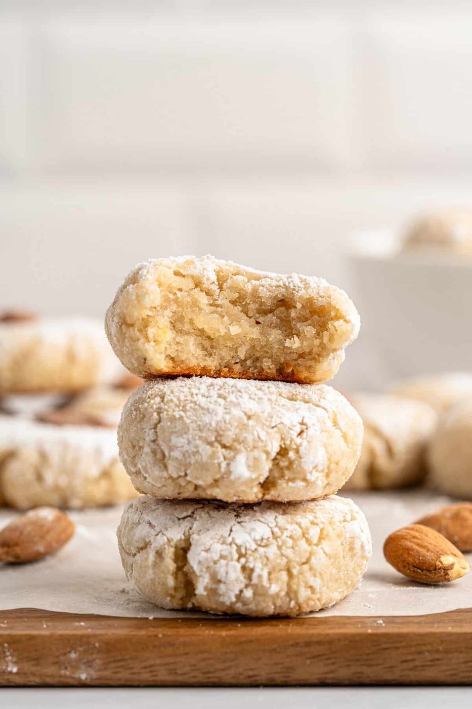 A stack of three powdered Italian Amaretti Cookies on a wooden surface. The top cookie has a bite taken out, revealing a soft interior. Whole almonds are scattered around, with more cookies blurred in the background.