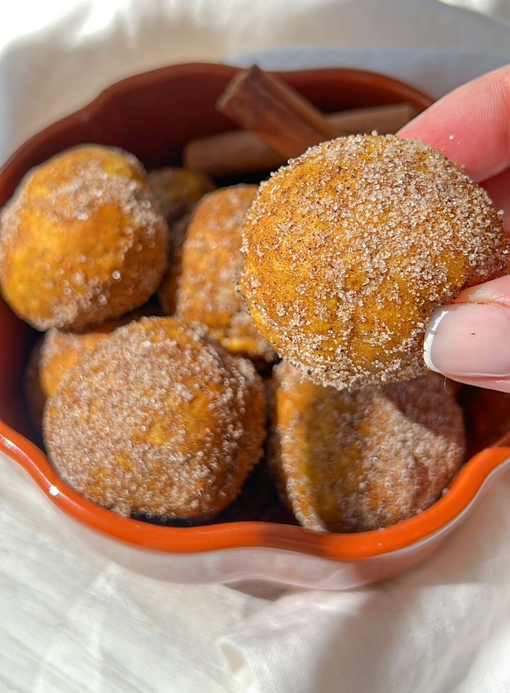 A bowl of cinnamon sugar-coated pumpkin donut holes. A hand holds one donut hole above the bowl. The background has a soft white cloth, and a few cinnamon sticks are visible in the bowl among the donut holes.