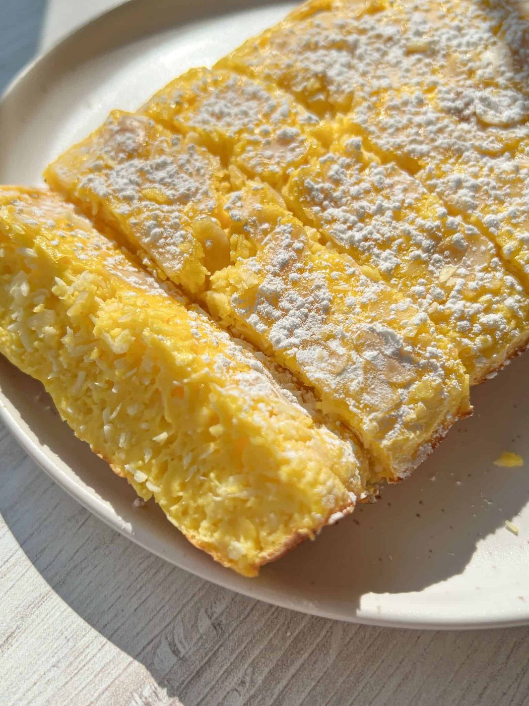 A close-up of a sliced 4 Ingredient Yogurt Cake dusted with powdered sugar on a beige plate. The texture appears moist and fluffy, with visible coconut flakes inside. Sunlight highlights the surface.