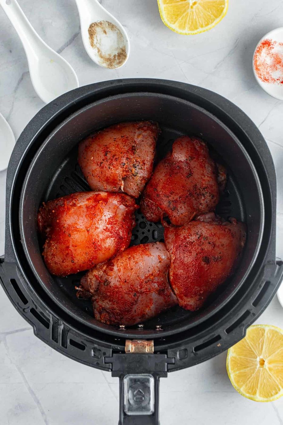 Air fryer containing seasoned and cooked chicken thighs. Slices of lemon and white spoons with spices are nearby on a marble surface.
