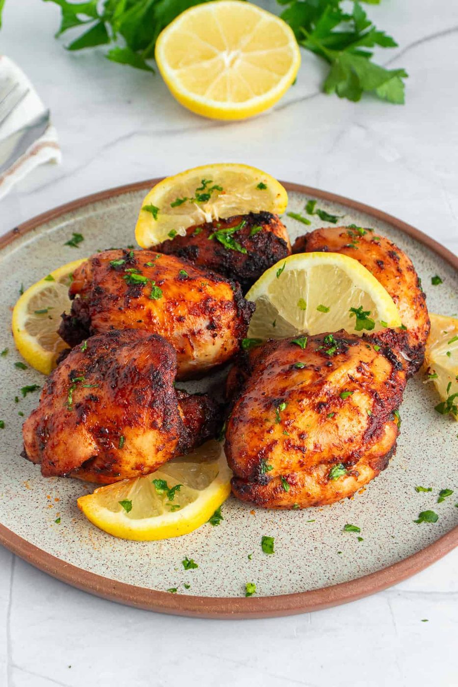 A plate of grilled chicken thighs garnished with lemon slices and fresh parsley. The chicken is browned and slightly charred, suggesting a flavorful, savory seasoning. In the background, there are cut lemons and parsley leaves.