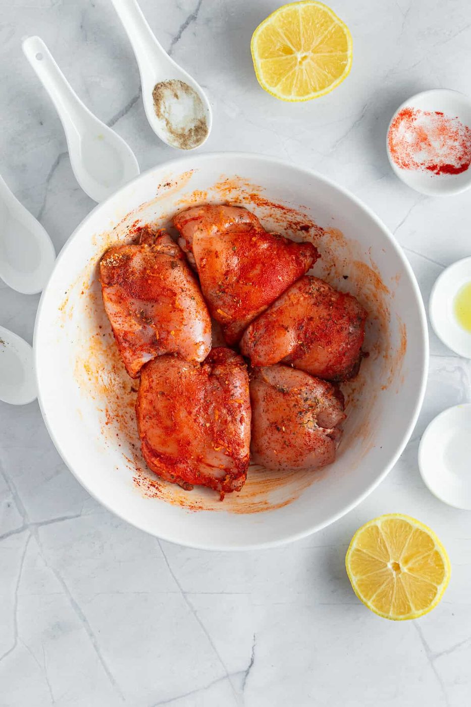 A white bowl contains raw chicken pieces marinated with spices. The bowl is surrounded by halved lemons and small white bowls filled with various spices on a marble surface. White measuring spoons also lie nearby.