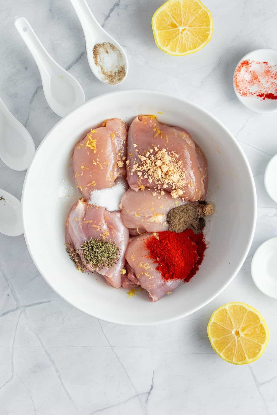 A white bowl filled with raw chicken pieces seasoned with spices, lemon zest, and herbs is surrounded by halved lemons and white spoons containing additional spices on a marble countertop.