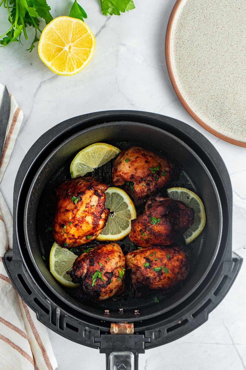 A top view of an air fryer basket containing four cooked chicken thighs garnished with lemon slices and herbs. A halved lemon, parsley, and a plate are placed nearby on the marble surface.