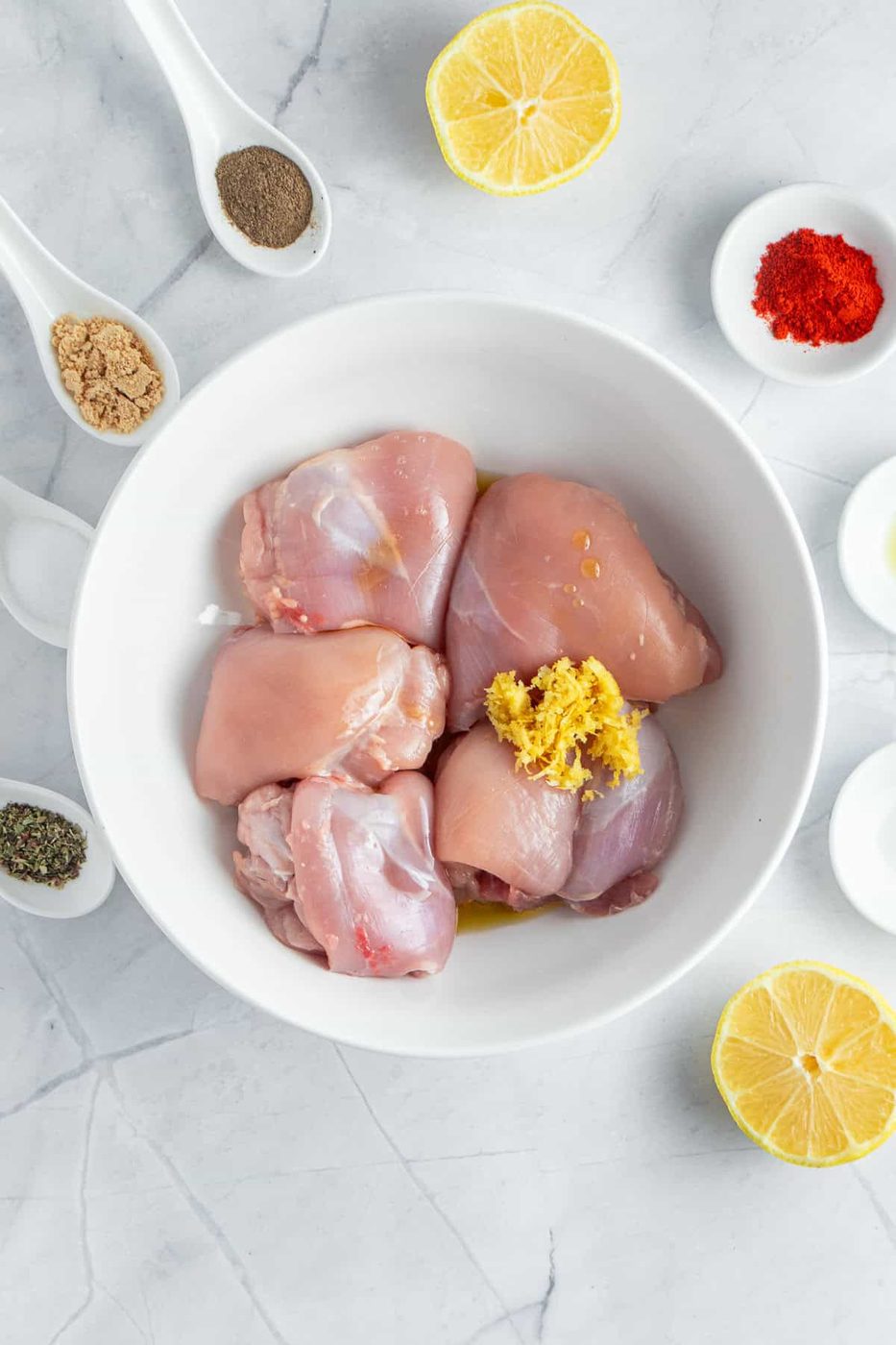 Chicken thighs in a white bowl with grated ginger on top, surrounded by lemon halves and small bowls containing ground spices, black pepper, salt, and red paprika, on a marble surface.