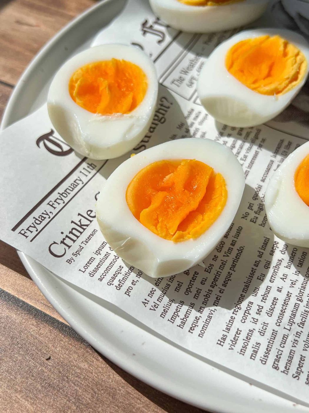Close-up of halved soft-boiled eggs with bright orange yolks on a white plate lined with a newspaper-style paper, sitting on a wooden surface.