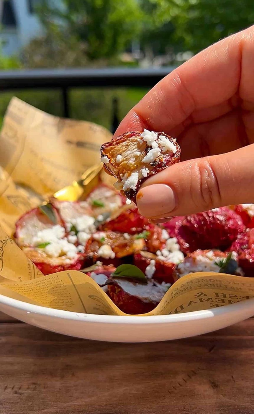 A hand holds a slice of roasted beet topped with crumbled cheese and herbs above a bowl lined with printed paper, filled with more roasted beet slices and garnishes.