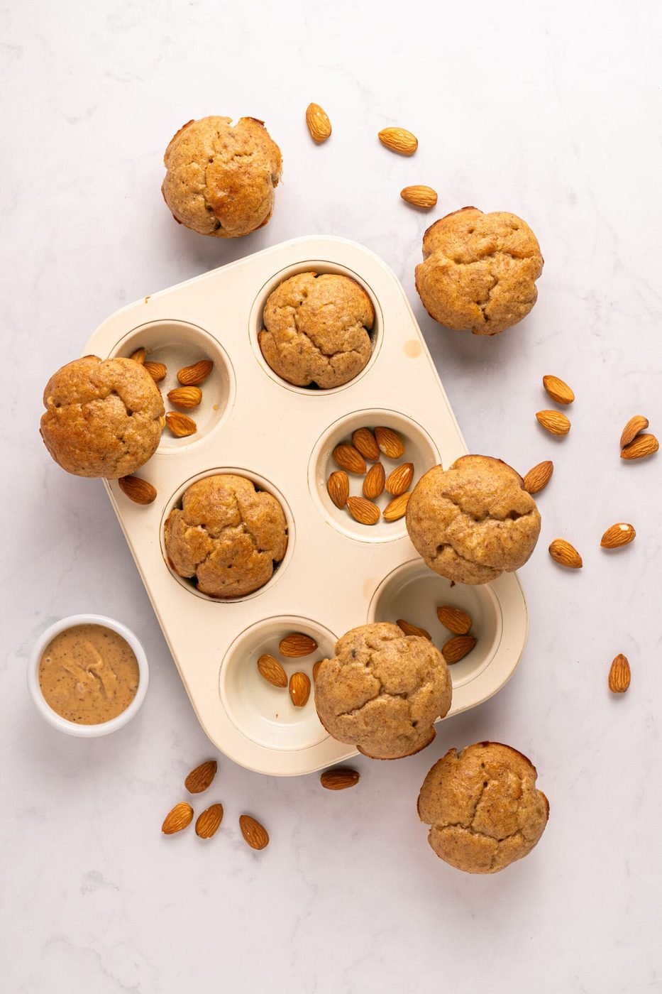 A white muffin tray with six almond muffins, surrounded by scattered whole almonds and a small bowl of almond butter on a white marble surface.