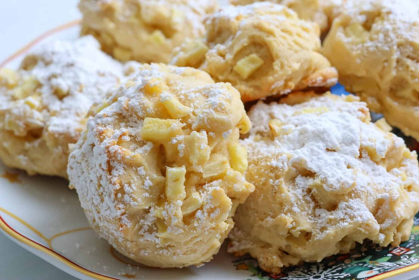 Close-up of several golden Apple Fritter Cookies sprinkled with powdered sugar, arranged on a decorative plate with a colorful pattern in the corner. The cookies have a chunky texture and visible pieces of apple.
