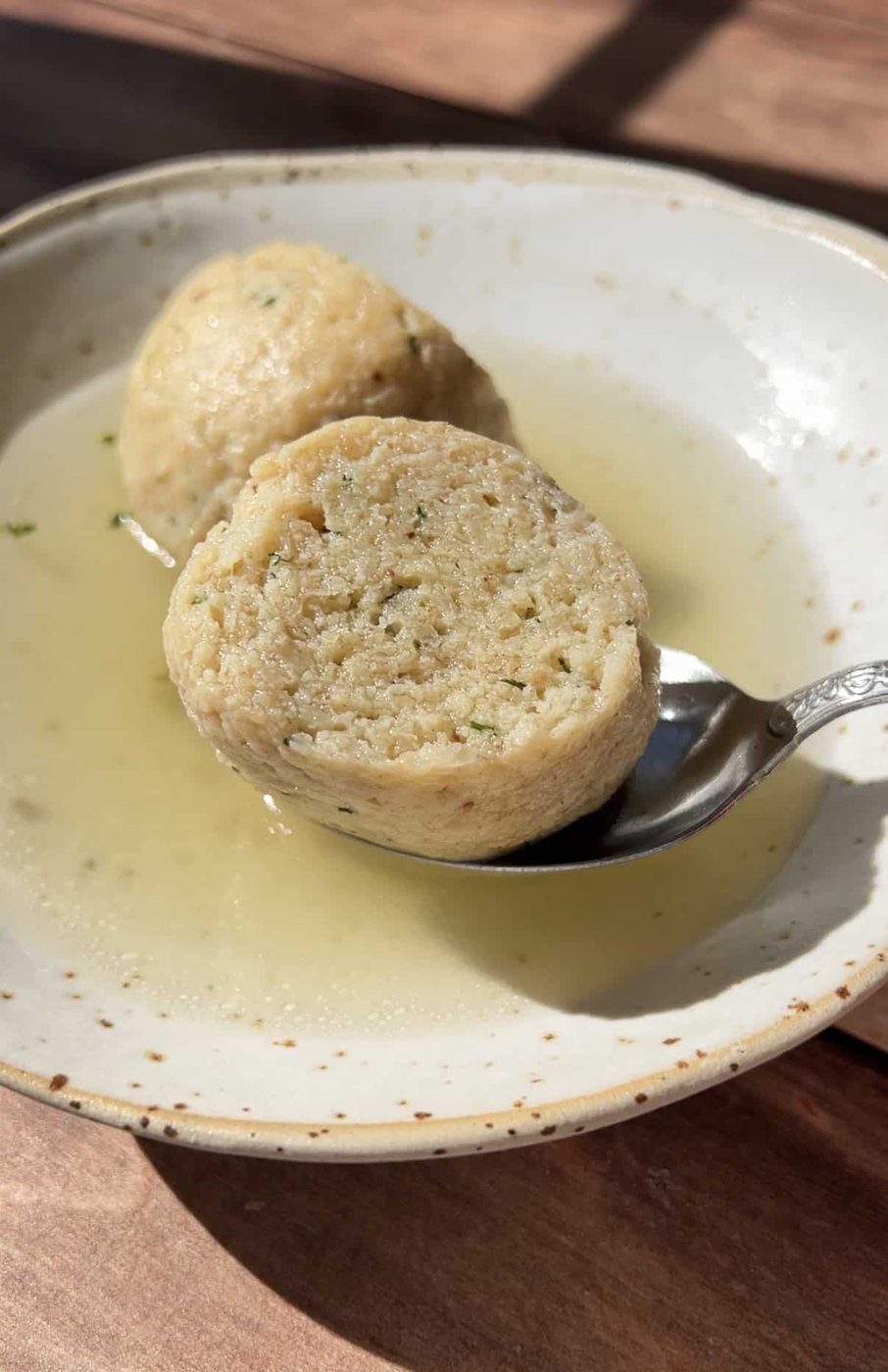 A close-up of a bowl with clear broth and a sliced bread dumpling, one half resting on a spoon. The dumpling appears soft with visible herbs, and sunlight highlights the textures.