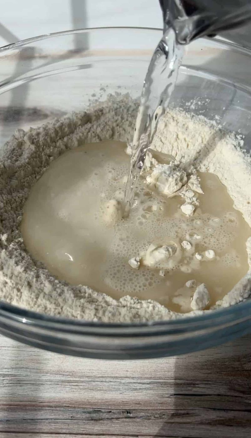 Water being poured into a glass bowl filled with flour, creating a well in the center. The scene is set on a light wooden surface, capturing the moment before mixing ingredients.