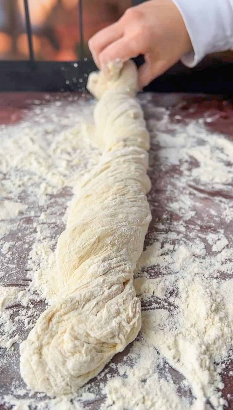 A hand is seen twisting a long, flour-dusted piece of dough on a floured wooden surface, preparing it for baking. The dough appears soft and fresh, with visible folds and texture.