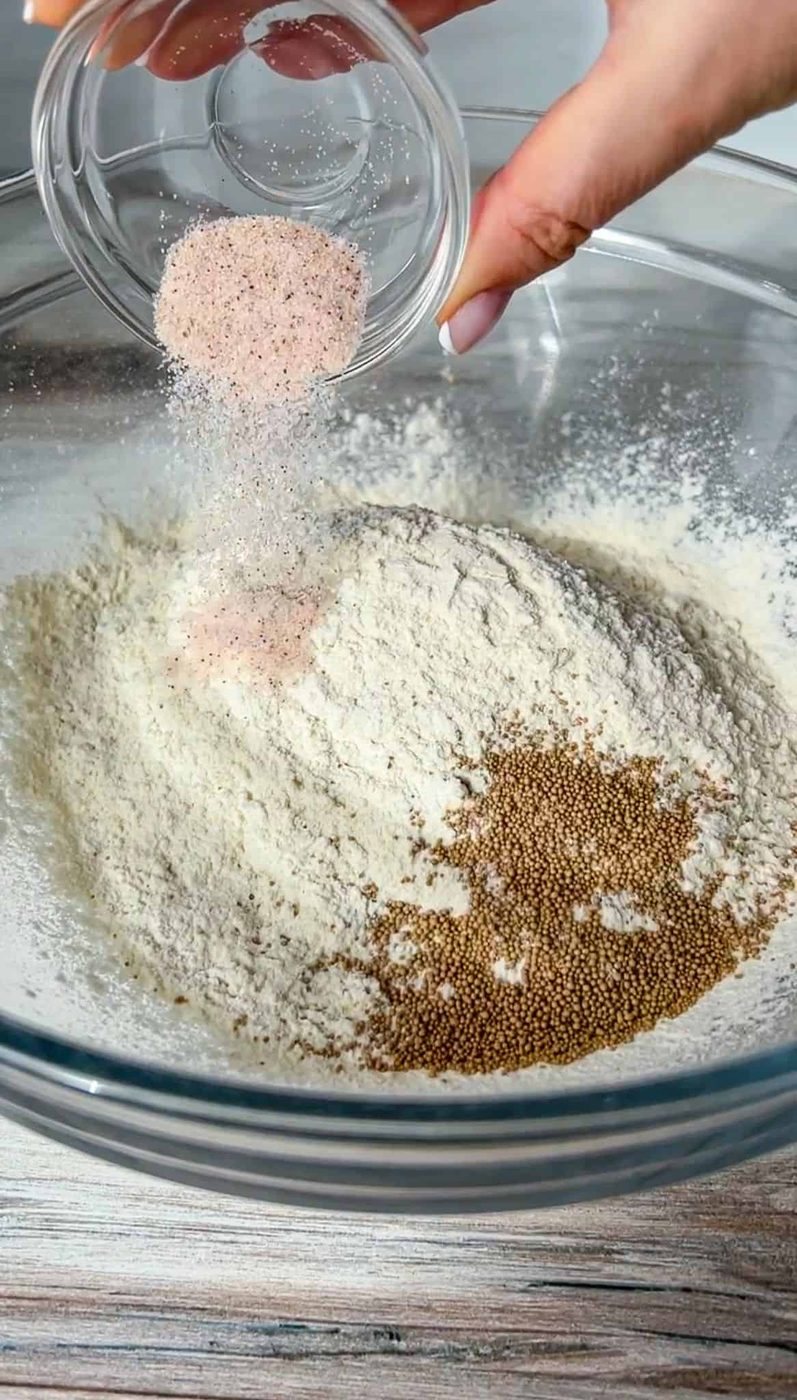 A hand pours pink salt and other seasonings from a small glass bowl into a large clear bowl containing flour and small brown seeds, likely yeast, on a wooden surface.