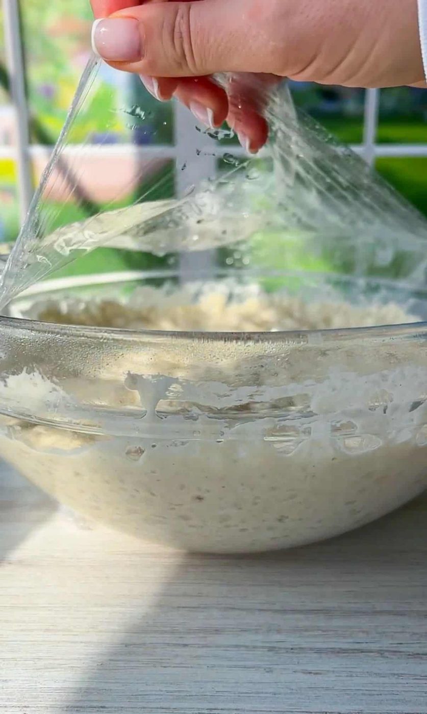 A hand is lifting plastic wrap off a clear glass bowl containing dough. The sunlight is illuminating the dough, highlighting its texture. The background shows a window with a blurred view of greenery outside.